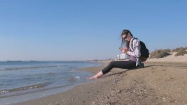 Gezgin kız kumlu deniz kumsalda oturuyor ve cep telefonunda bir mesaj yazarak bir sırt çantası ile.