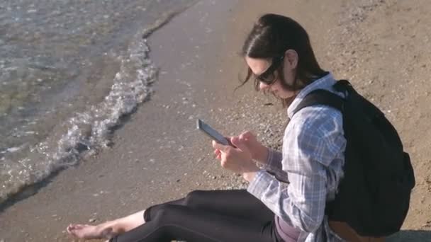 Menina viajante com uma mochila sentada em uma praia de areia e digitando uma mensagem no telefone celular . — Vídeo de Stock