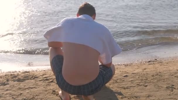 El hombre está sentado en la playa, cubriéndose los hombros con una toalla del sol . — Vídeo de stock