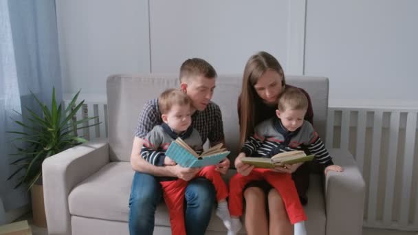 家族のお母さん、お父さんと 2 つの双子の兄弟は、ソファに座って本を読みます。家族の読書の時間. — ストック動画