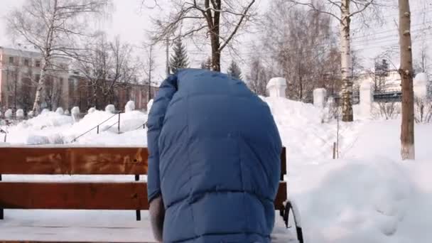 Женщина убирает скамейку со снега и сидит на скамейке в зимнем городском парке в течение дня в снежную погоду с падающим снегом . — стоковое видео