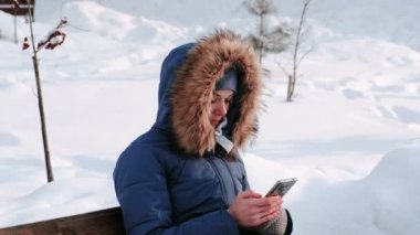 Tatlı kadın bankta oturan ve gün yağan kar ile karlı havalarda sırasında cep telefonu şehir parkında kış tarama.