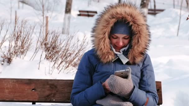 La mujer de primer plano está sentada en el banco y navegando por el teléfono móvil en el parque de invierno de la ciudad durante el día con clima nevado con nieve cayendo . — Vídeos de Stock