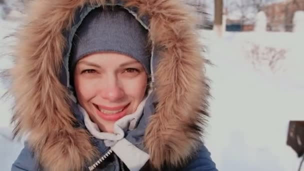 Young beautiful woman in fur hood talking on video connection and walking in winter city park in snowy day with falling snow. — Stock Video
