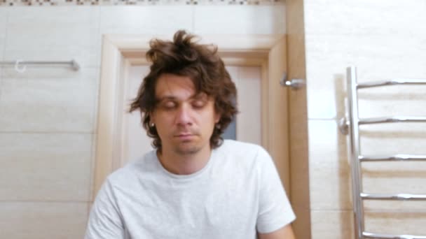 Sleepy shaggy joven se mira al espejo en el baño por la mañana y peina el pelo largo con las manos . — Vídeos de Stock