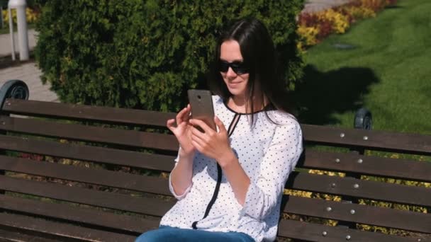 Jonge vrouw brunette typt een bericht op mobiele telefoon zittend op het bankje in park. — Stockvideo