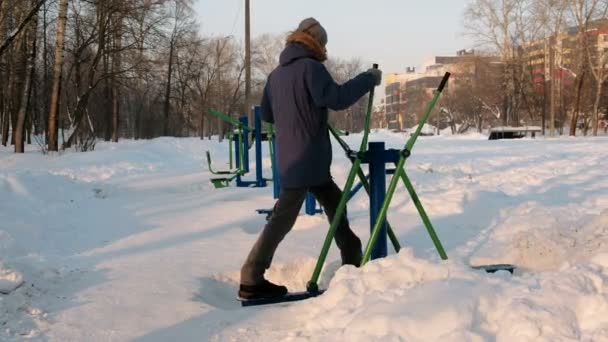 Mann in blauer Daunenjacke mit Kapuze sitzt in einem Winterstadtpark im Skisimulator. zurück. — Stockvideo