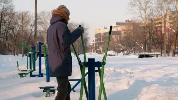 Mann in blauer Daunenjacke mit Kapuze sitzt in einem Winterstadtpark im Skisimulator. zurück. — Stockvideo