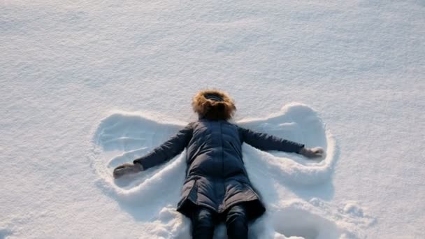 Kvinna i blå dunjacka med en päls huva gör en snöängel. — Stockvideo
