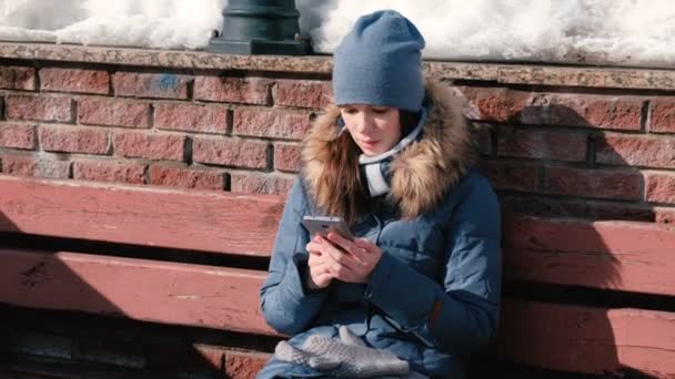 Mujer está leyendo un libro en el teléfono móvil sentado en el parque de invierno . — Vídeos de Stock