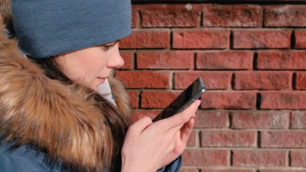 Mulher está navegando páginas da internet no telefone móvel sentado no parque de inverno. Vista lateral do rosto close-up . — Vídeo de Stock