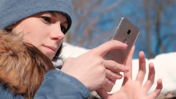 Vrouw is surfen op internetpagina's op mobiele telefoon zitten in winter park. Closeup gezicht — Stockvideo