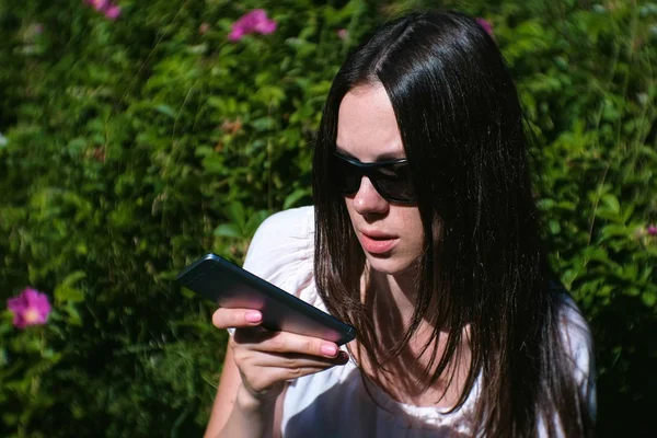 Hermosa morena graba un mensaje de voz en su teléfono móvil mientras está sentada en el parque en un día soleado . — Foto de Stock