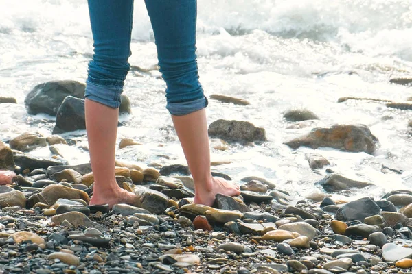 女性は、波のように石のビーチに立っている.クローズ アップ ジーンズの足. — ストック写真