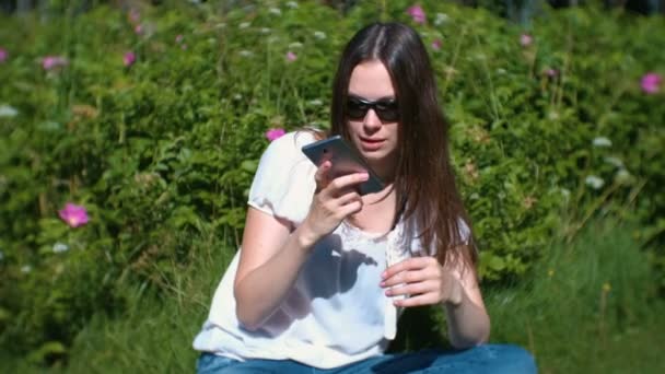 Belle femme brune enregistre un message vocal sur son téléphone portable alors qu'elle est assise dans le parc par une journée ensoleillée . — Video