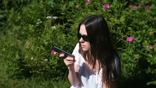 Beautiful brunette woman records a voice message on her mobile phone while sitting in the park on a Sunny day. — Stock Video