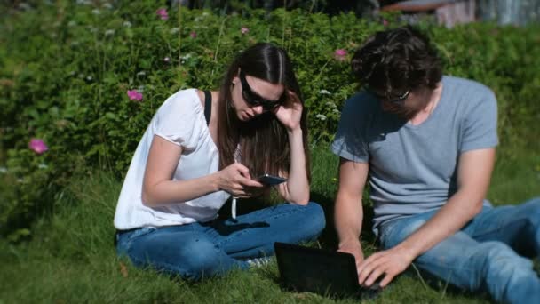 Young couple man and woman are working together on mobile and laptop sitting in park. — Stock Video