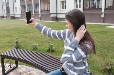 Güzel esmer bankta otururken rüzgarda cep telefonunda bir selfie almaya çalışıyor.