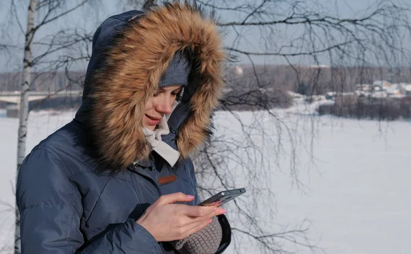 Close-up mulher em azul jaqueta com capuz de pele escreve mensagens em seu celular em um parque de inverno — Fotografia de Stock