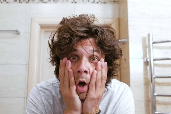 Sleepy shaggy young man looks at the mirror in bathroom in the morning and surprised. — Stock Photo, Image