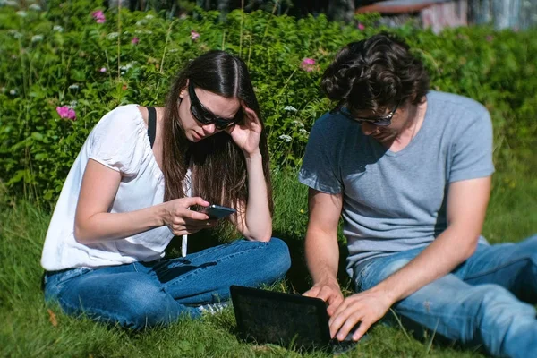 Pareja joven hombre y mujer están trabajando juntos en el móvil y portátil sentado en el parque . — Foto de Stock