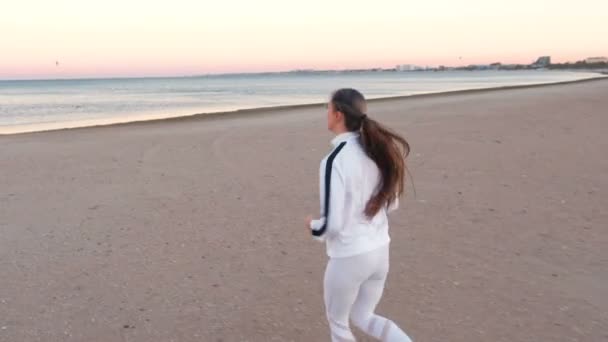 Młoda kobieta jest jogging na piaszczystej plaży nad morzem na wschód słońca w jesieni, widok z boku. — Wideo stockowe