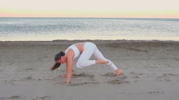 Young woman is doing yoga Adho mukha shvanasana with raised foot on the sand beach at sunrise. — Stock Video