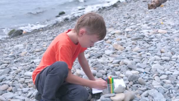 Ragazzo sta dipingendo disegna con pennarelli mentre seduto su una spiaggia di pietra sulla riva del mare . — Video Stock