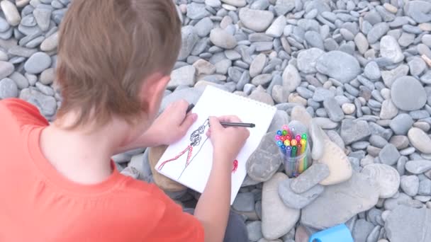 Jongen trekt met markeringen zittend op een stenen strand aan de oever van de zee. Achteraanzicht. — Stockvideo