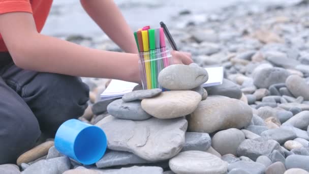 Close-up jongens handen is schilderen met markeringen zittend op een stenen strand aan de oever van de zee. — Stockvideo