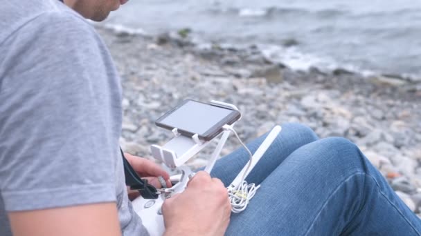 Adam bir dron taş bir plajda denizin kenarında oturan bir uzaktan kumanda ile kontrol eder. Yakın çekim yakışıklı. — Stok video