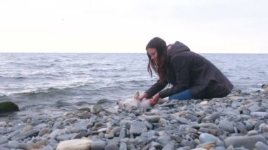 Genç kadın denizin kenarında taş sahilde beyaz köpeğiyle oturuyor.