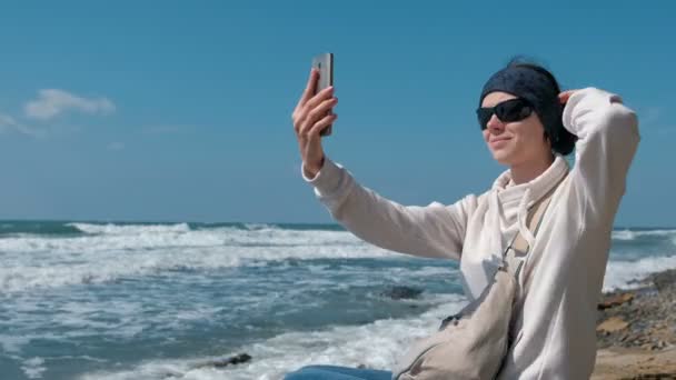 Жінка змушує селфі сидіти біля моря на кам'яному пляжі восени . — стокове відео