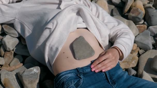 Piedra en el vientre de las chicas. Chica irreconocible acostada en una playa de piedra en el otoño . — Vídeos de Stock