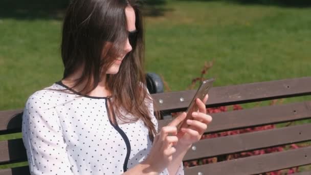 Ung kvinna brunett talar videochatt på mobiltelefon sitter på bänken i parken. — Stockvideo