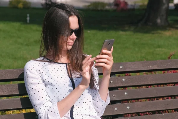 Ung kvinnelig brunette skriver en beskjed på mobilen sittende på benken i parken. . – stockfoto