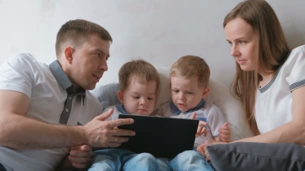 Rodina s tabletem. Máma, táta a dva syny dvojčata batolata pohledu karikatura na tablet ležící na posteli. — Stock video