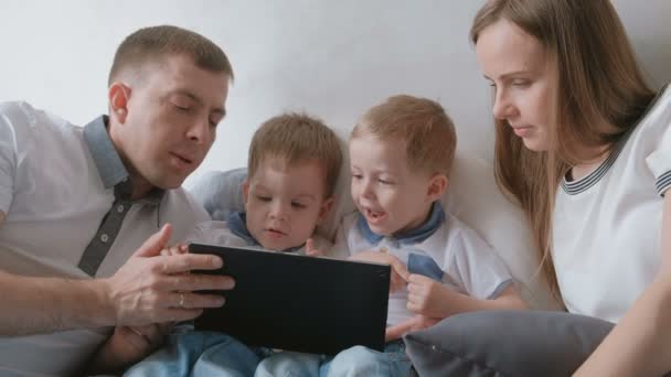 Famille avec tablette. Maman, papa et deux fils jumeaux tout-petits regardant dessin animé à la tablette couché sur le lit . — Video