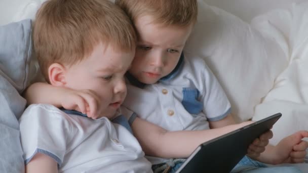 Děti s tabletem. Dva kluky dvojčata batolata hledá karikatury na tablet ležící na posteli. — Stock video