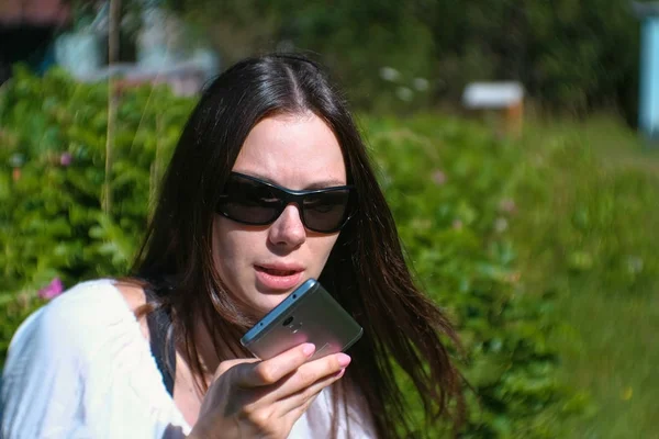 Hermosa morena graba un mensaje de voz en su teléfono móvil mientras está sentada en el parque en un día soleado . — Foto de Stock