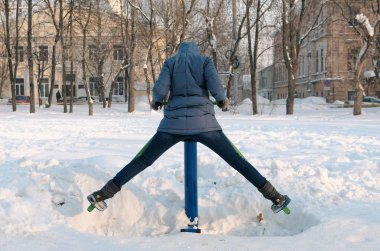 Kadın mavi ceket bir kukuleta ile aşağı bacak egzersizleri simülatör şehir arka görünümünde bir kış parkta yapıyor