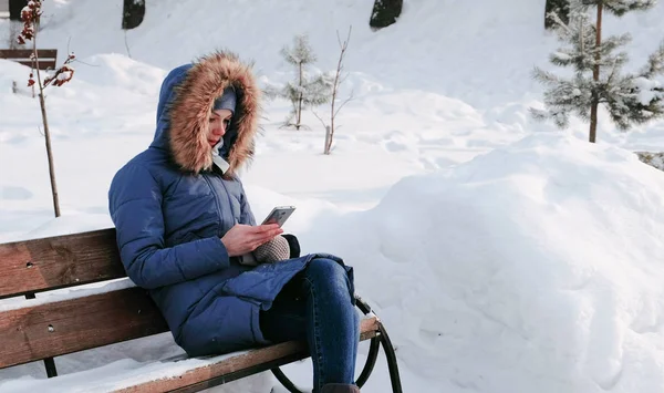 Женщина сидит на скамейке и просматривает мобильный телефон в зимнем парке в городе в солнечный день . — стоковое фото