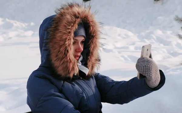 Jovem mulher bonita feliz chamada vídeo conversando no parque de inverno na cidade em dia nevado com neve caindo. Vista lateral . — Fotografia de Stock