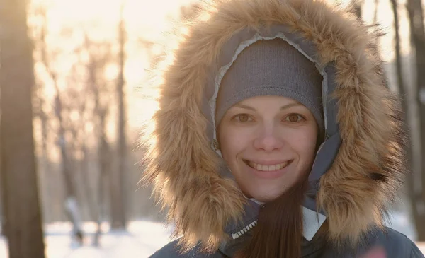 Крупним планом жінка в хутряному капюшоні дивиться в камеру і посміхається в зимовому сонячному парку . — стокове фото