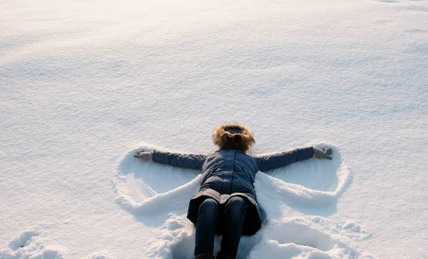 Mulher de casaco azul para baixo com um capuz de pele faz um anjo da neve . — Fotografia de Stock