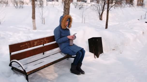 Mujer está sentada en el banco y el teléfono móvil de navegación en el parque de invierno en la ciudad durante el día en el tiempo nevado con nieve cayendo . — Vídeo de stock