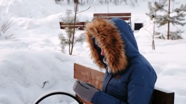 Closeup view of woman is sitting on bench and browsing mobile phone in winter city park during the day in snowy weather with falling snow. — Stock Video