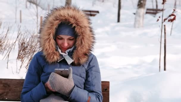 La mujer de primer plano está sentada en el banco y navegando por el teléfono móvil en el parque de invierno de la ciudad durante el día con clima nevado con nieve cayendo . — Vídeos de Stock