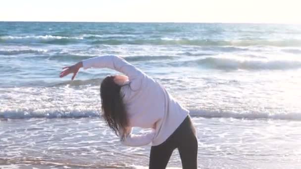 Frau dehnt sich und macht Yoga im Stehen am Sandstrand mit Wellen, Rückansicht. — Stockvideo