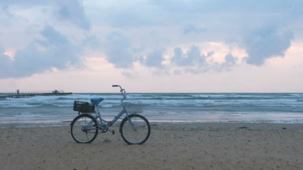 Old bicycle on the beach at sunset with storm sea and foam waves background and pier with walking people. — Stock Video